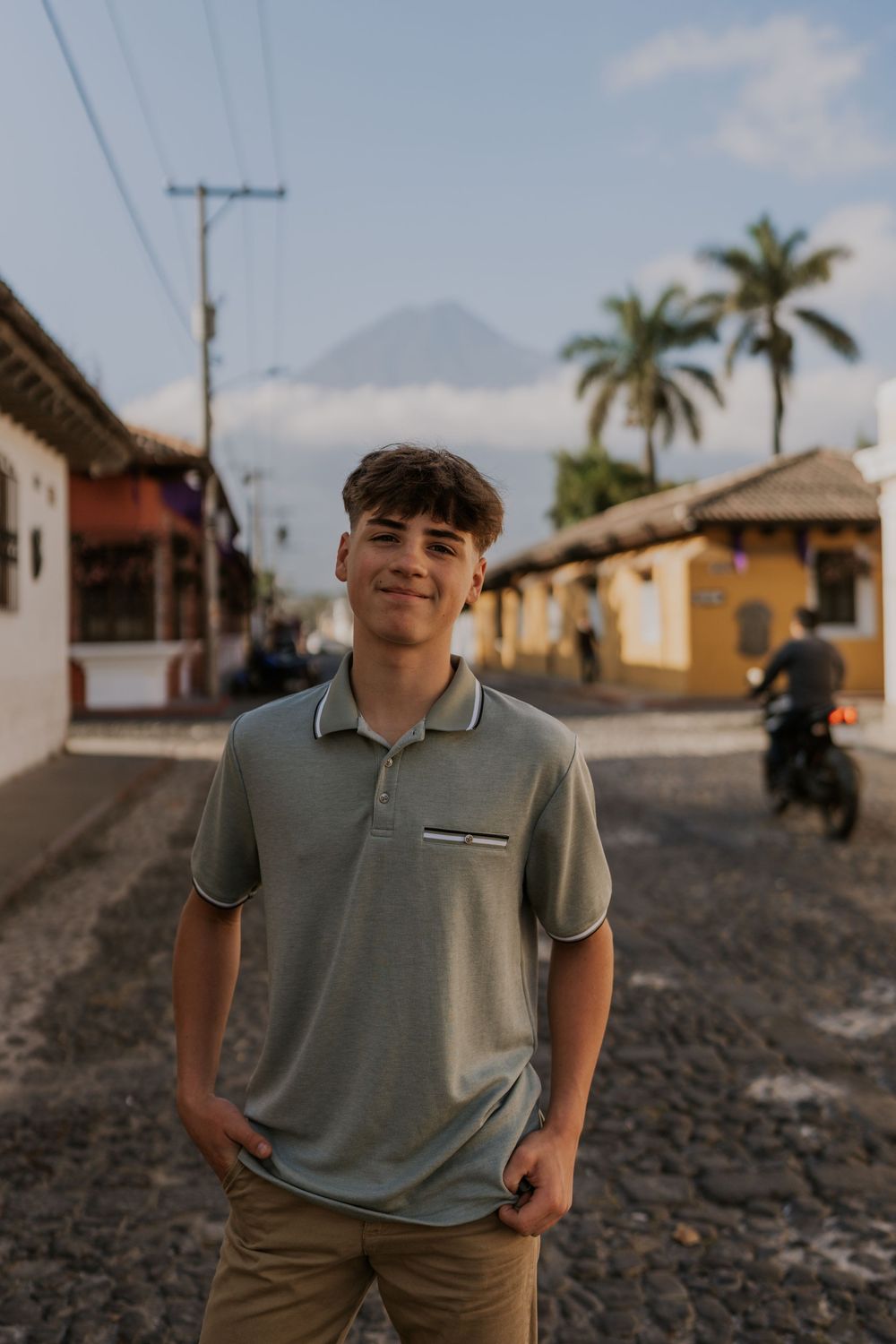 A person in a light gray polo shirt poses against a scenic coastal backdrop with cobblestone streets and palm trees.