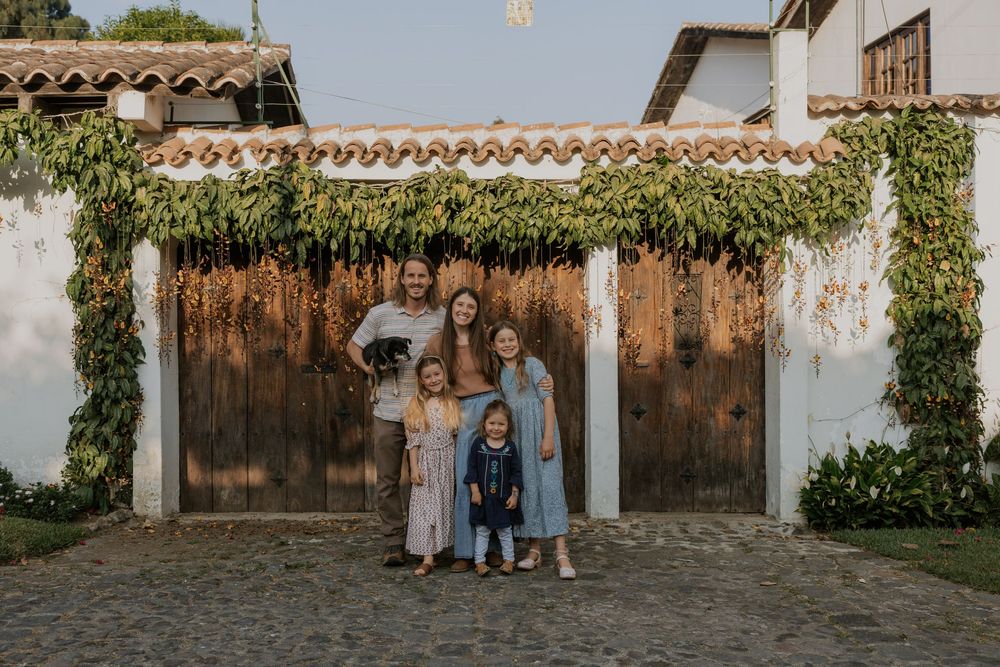 Family members in coordinated blue outfits pose against a rustic white stucco wall with wooden doors and climbing vines.