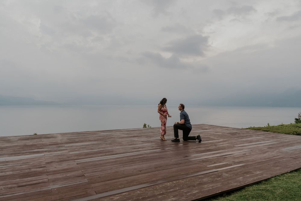 Man proposes on knees to woman on wooden deck overlooking calm ocean under cloudy sky.
