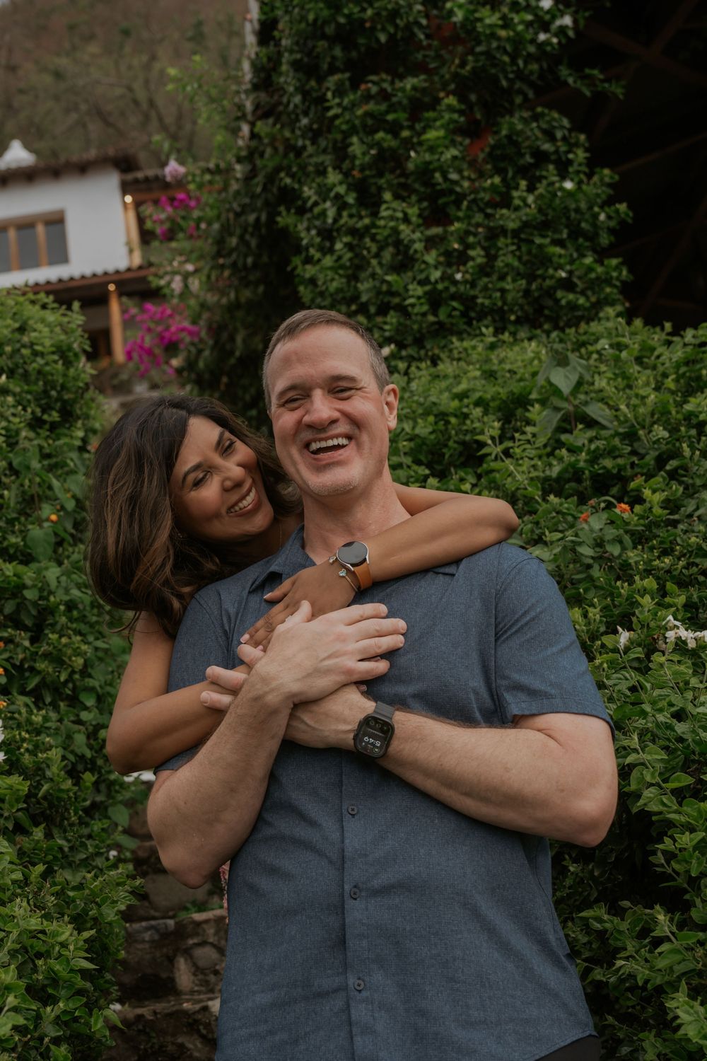 A happy couple embraces outdoors, surrounded by lush green foliage and colorful flowers near a rustic building.