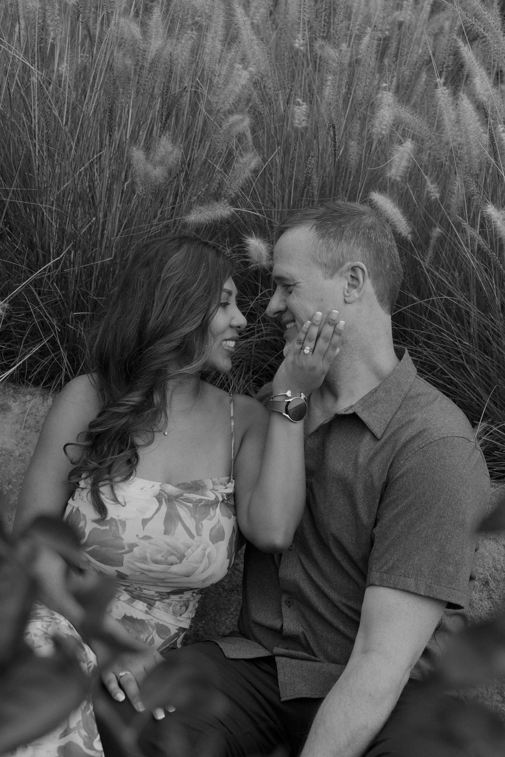 Black and white photo of a romantic couple sitting in tall grass, smiling at each other during an outdoor engagement session.