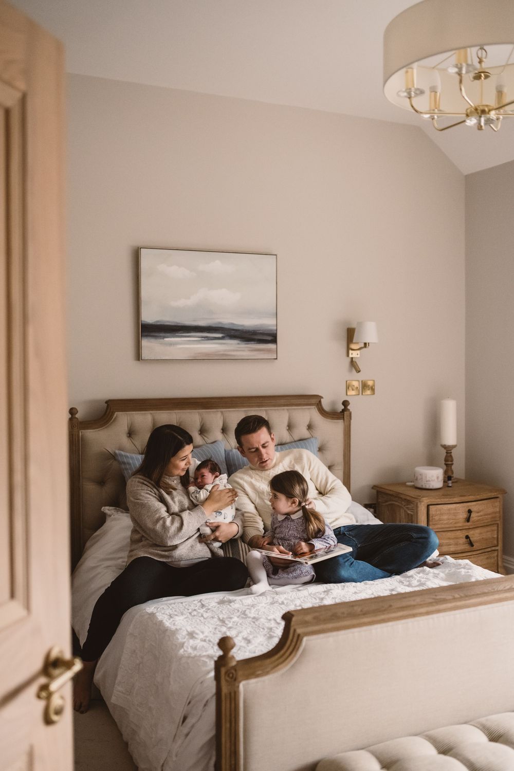 Family of four with a newborn baby and toddler snuggle together and reads in master bedroom with wooden furniture and beige walls.