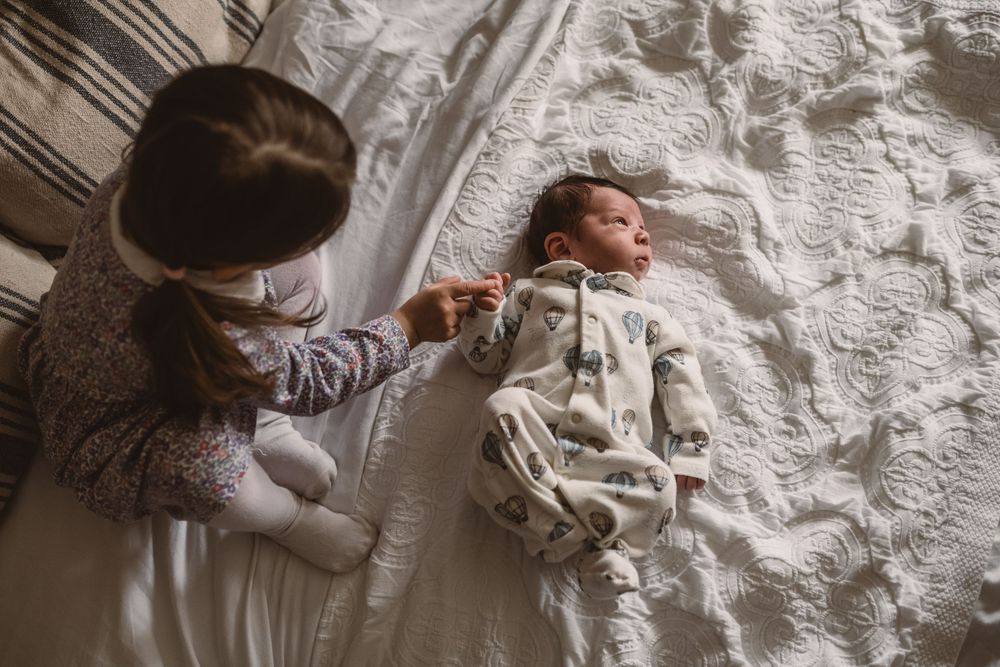 A toddler reading out to a newborn baby lying on while bedding.