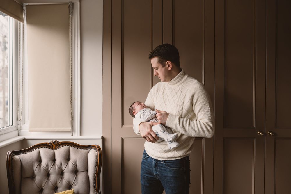 A father wearing a cream sweater holds a newborn baby while standing by a window beside a tufted chair.