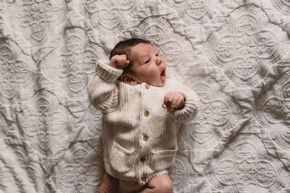 Newborn baby in white knit sweater lying on a textured cream-colored blanket.