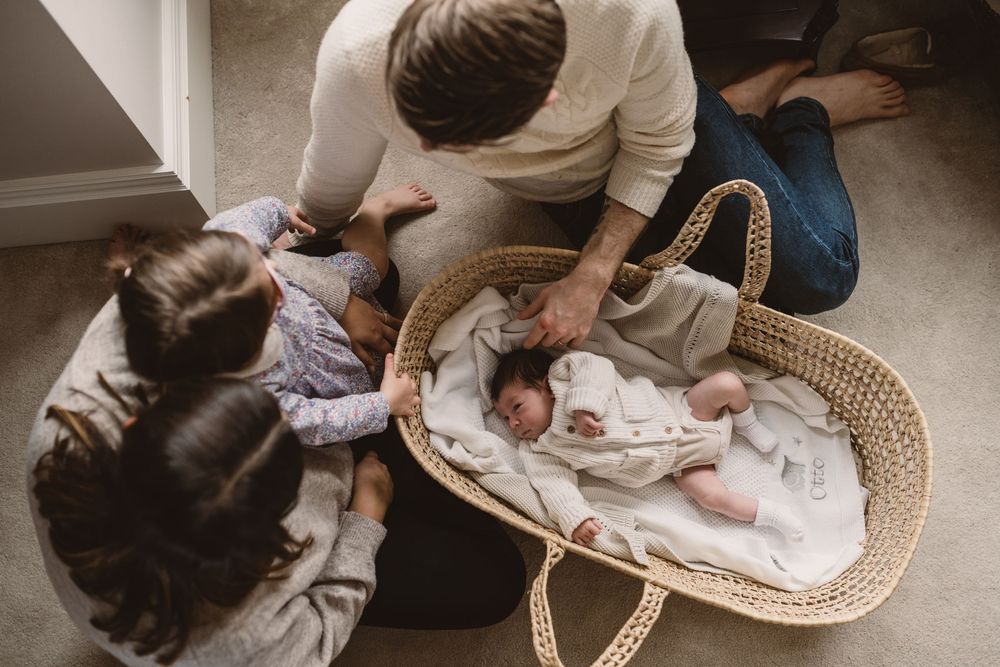 Several hands gently caress a sleeping newborn baby in a woven moses basket.