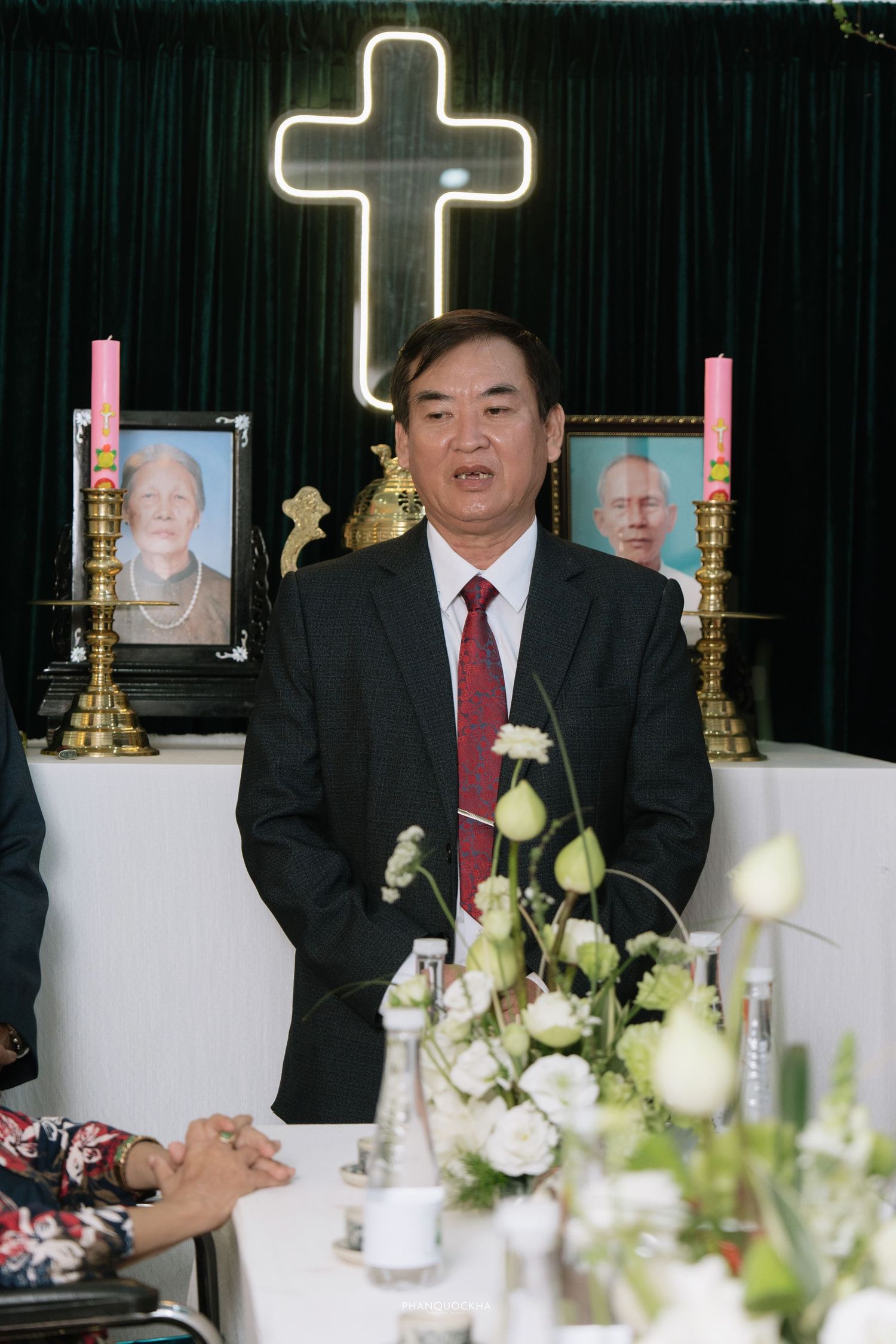 Man in black suit with red tie speaking at Vietnamese wedding altar decorated with cross and candles.