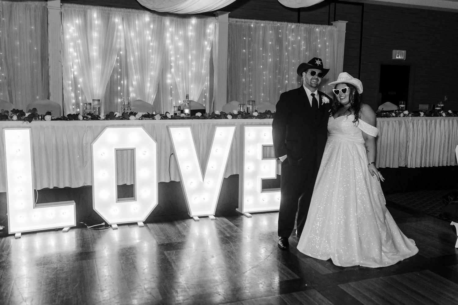 A black and white wedding reception photo showing a couple standing next to illuminated LOVE letters.