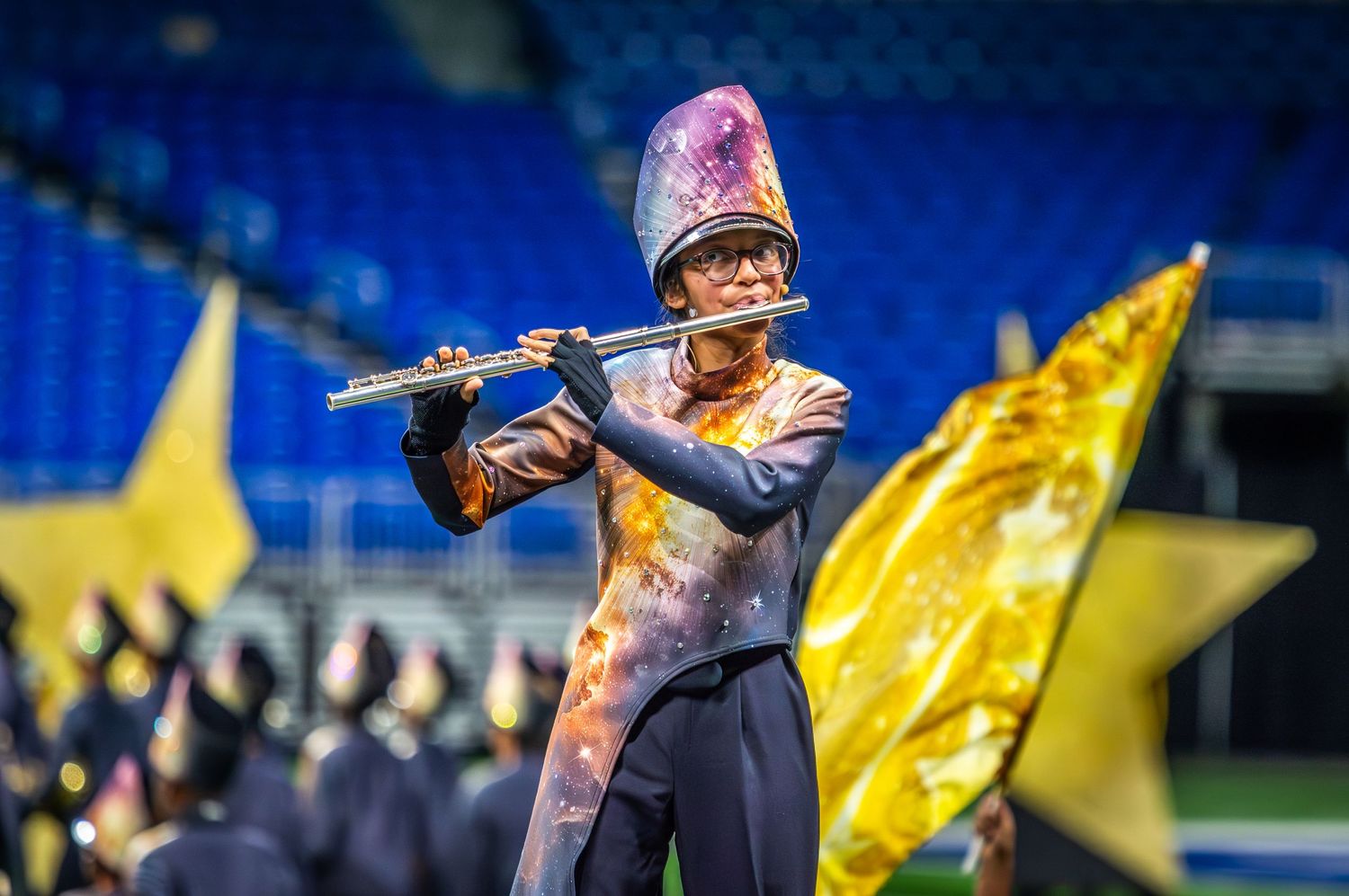 Marching band member plays flute in uniform with gold flags and blue stadium seats in background.