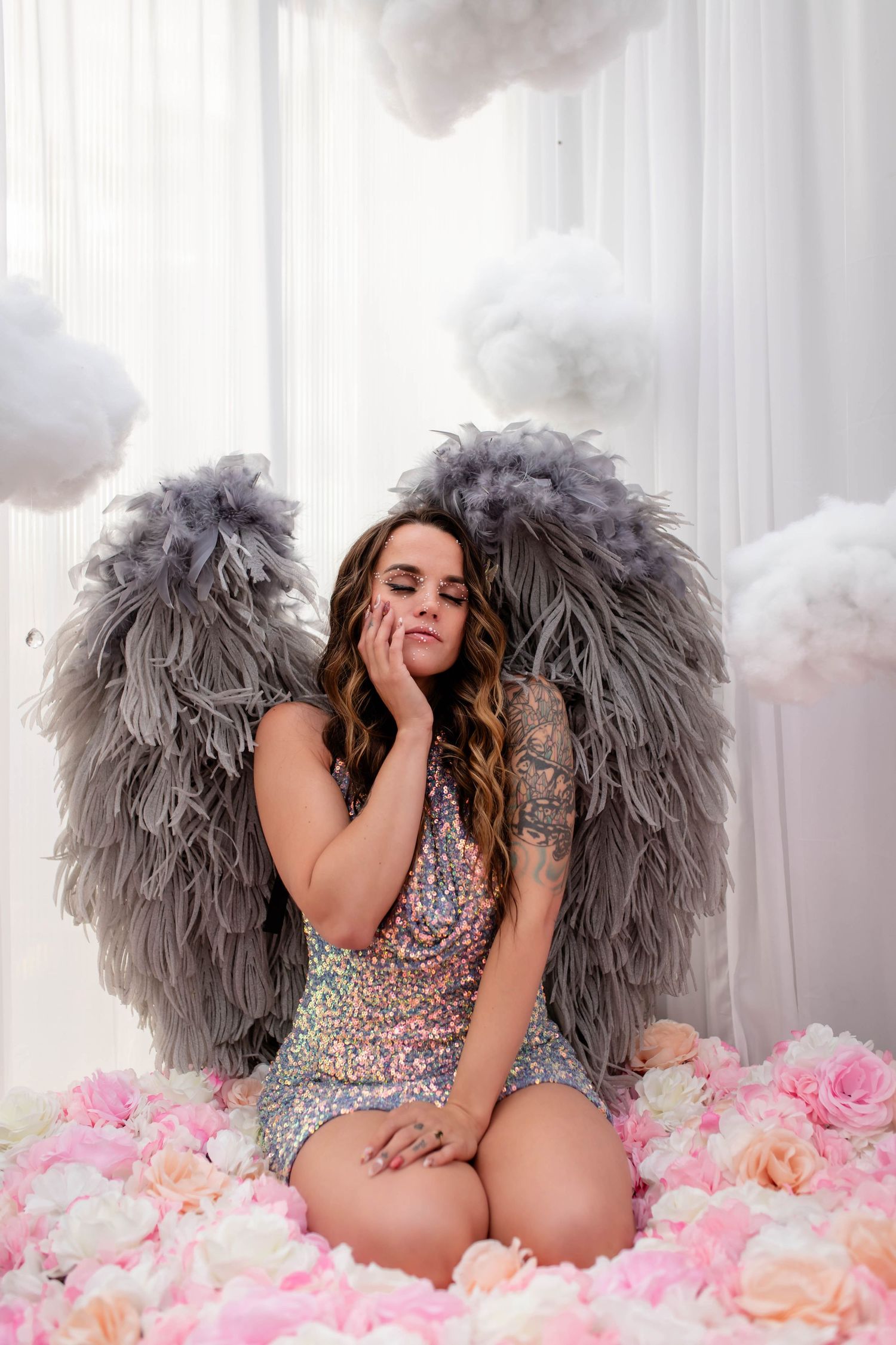 Dreamy studio portrait with grey feather wings and pink flower petals against white backdrop.