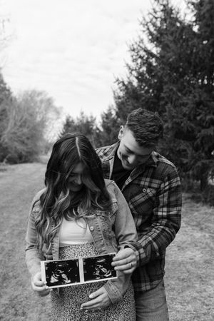 A couple sharing a joyful moment outdoors while holding ultrasound photos in a black and white photograph.