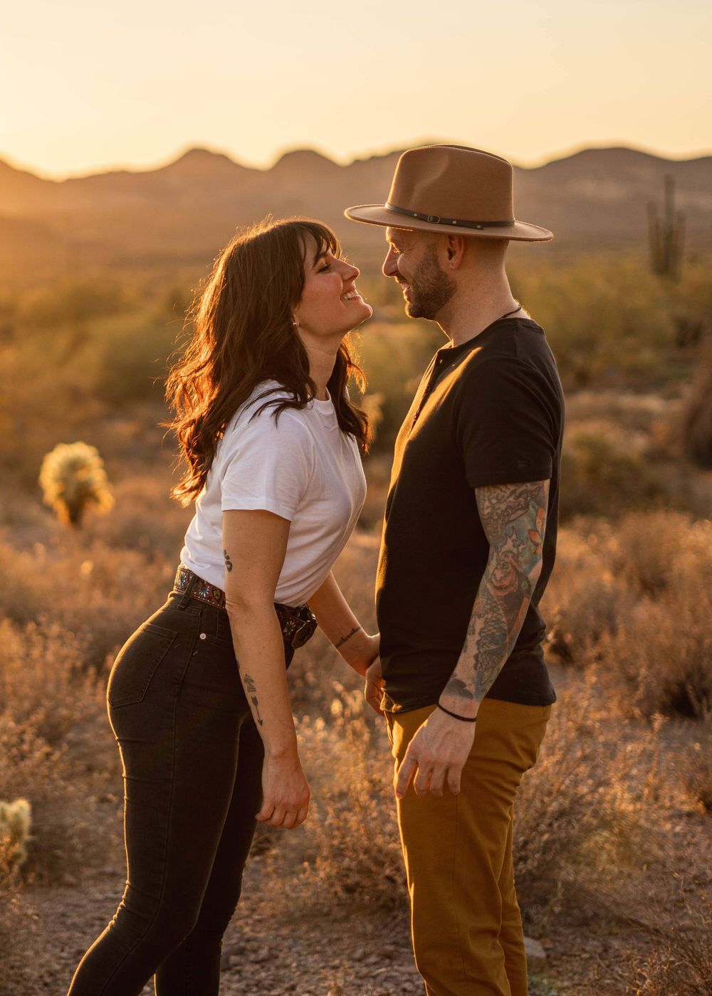 Couple sharing romantic moment in desert with one wearing brown hat during golden hour.