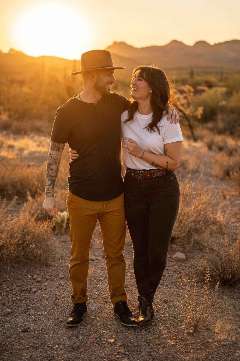 A couple walks together in a desert landscape during golden hour sunset, silhouetted against the warm glowing sky.