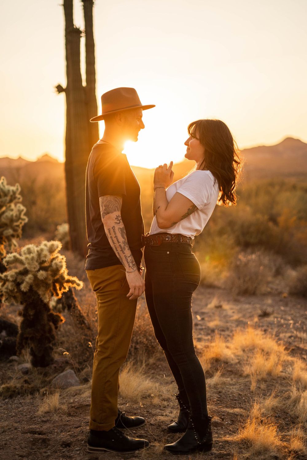 Silhouetted couple embraces in golden desert sunset light with cacti and desert brush in background.