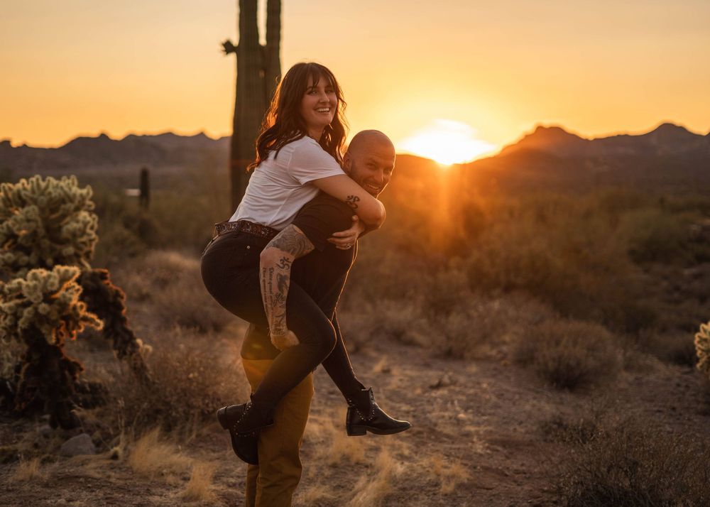 A couple embraces during golden hour in a desert landscape as the sun sets behind them creating a warm silhouette.