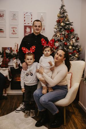 A family poses together near a decorated Christmas tree in a cozy living room with holiday decor on the walls.