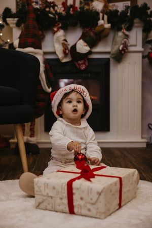 Baby in white outfit sits on floor with large wrapped Christmas present during holiday photoshoot.