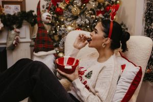 Person in white Christmas sweater enjoys holiday snacks while relaxing in cozy chair by decorated tree.