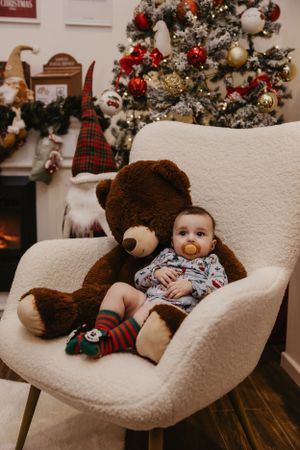 Baby sits with large teddy bear in white chair near Christmas tree wearing festive socks.