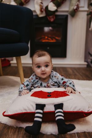 Baby lying on Santa pillow wearing holiday pajamas and striped socks near fireplace.