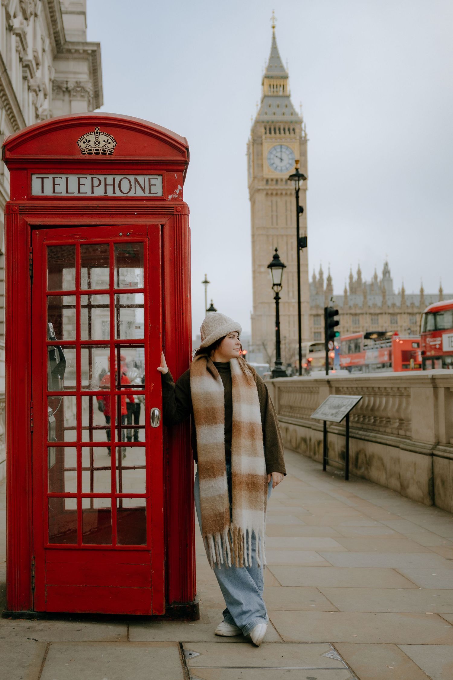 Woman in a beanie and plaid scarf poses beside a red British telephone box with Big Ben in the background in London.