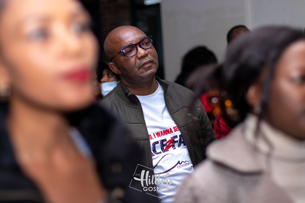 Person wearing glasses and a white t-shirt sits attentively in an audience during a live performance.