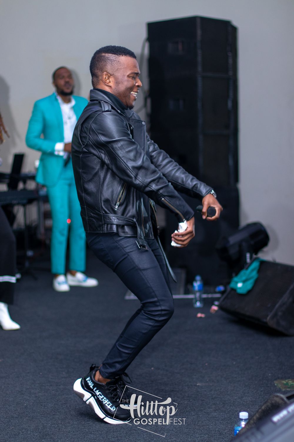 Takie Ndou in black leather jacket and jeans dances energetically on stage with Collin Maluleke in turquoise suit in background.
