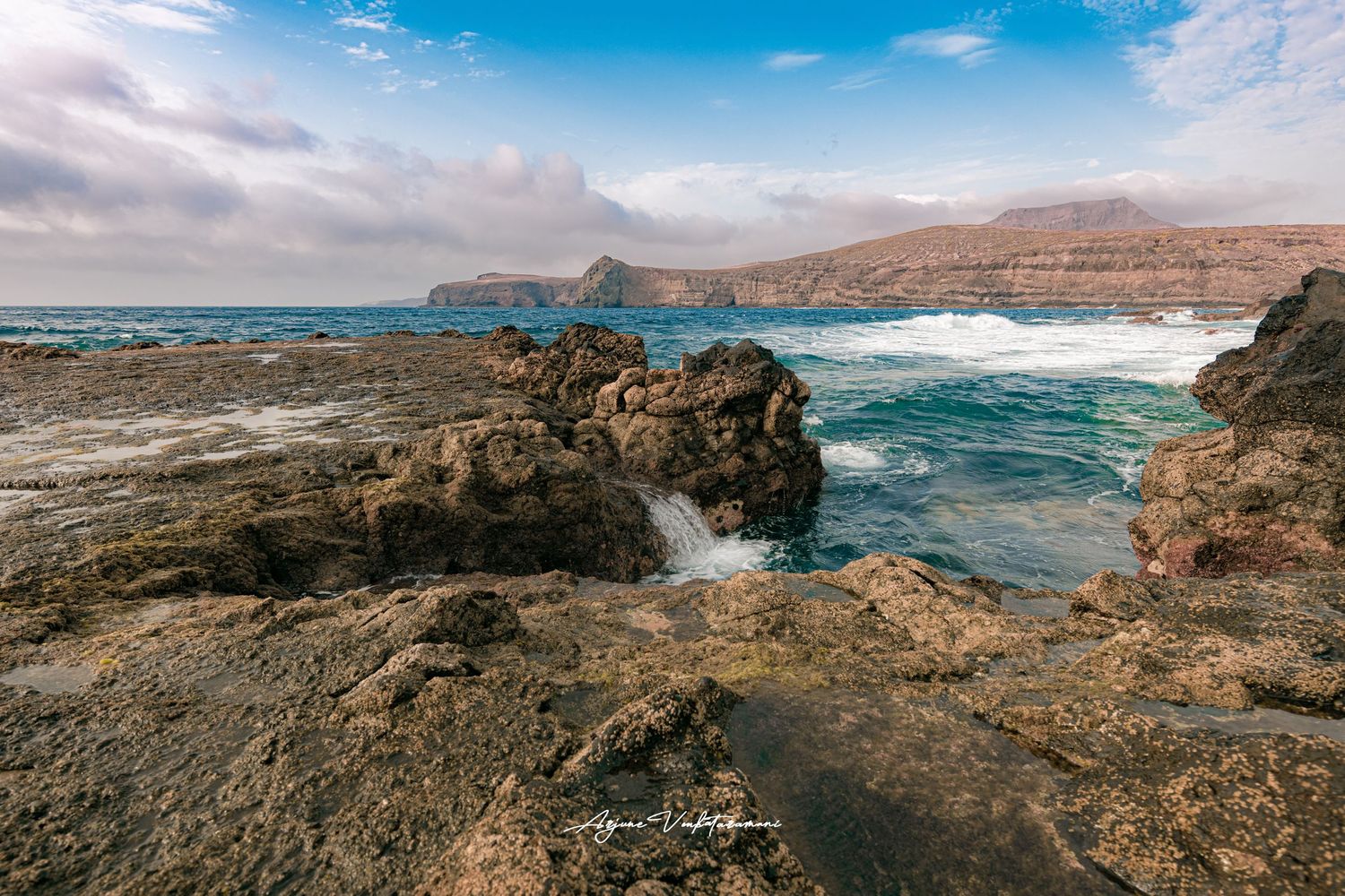 Rugged volcanic coastline with crashing waves and distant mountains along the shoreline under blue skies in Lanzarote, Spain.