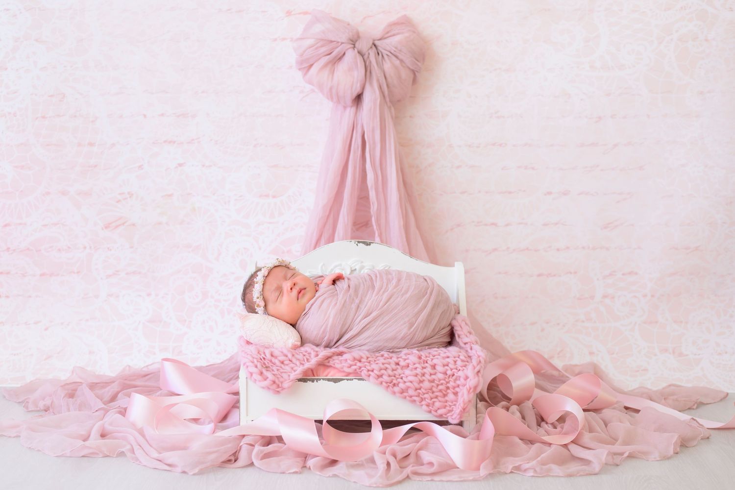 Peaceful newborn sleeps on white vintage bed with soft pink cushions beneath a matching pink bow canopy draped from above.