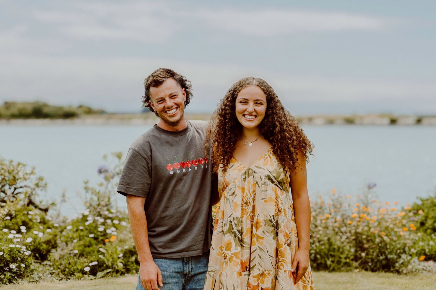 Couple poses together on coastal path with water and greenery in background.