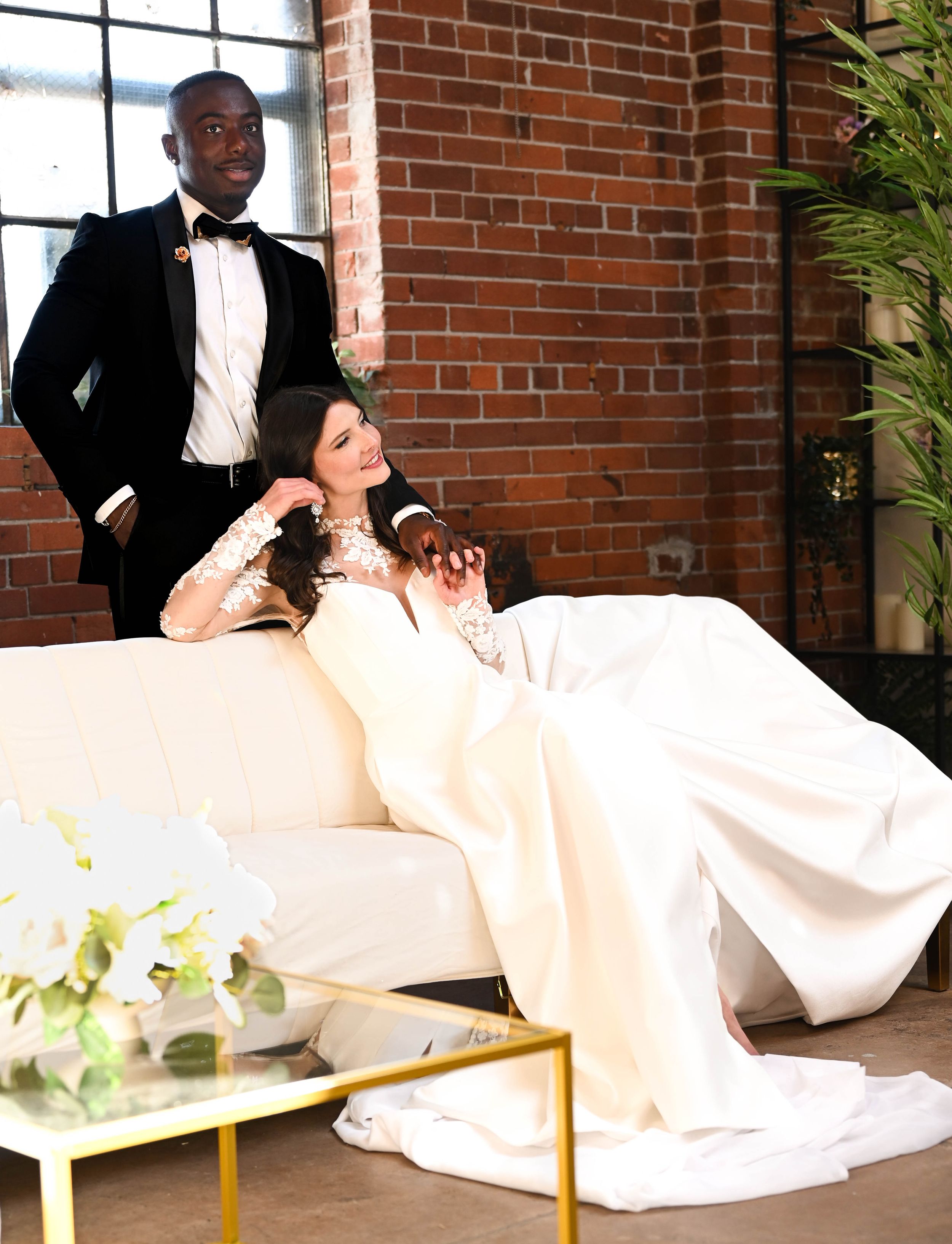 A wife sits on a couch in her wedding dress as her husband laces his fingers with hers as he stands behind her.