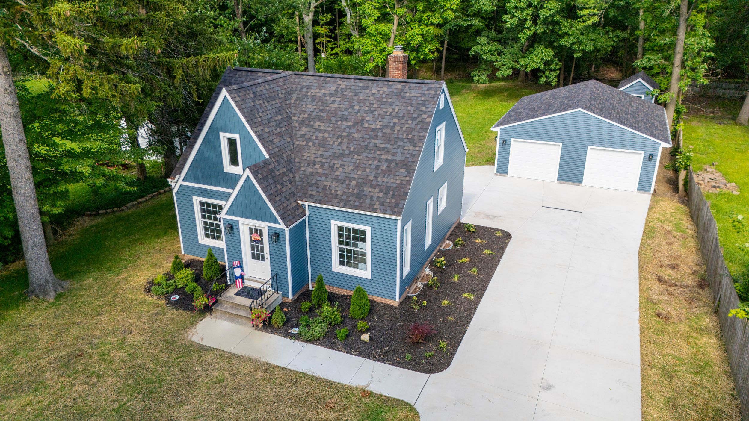 Aerial drone panorama of blue house with attached deck and shaded backyard surrounded by green trees.