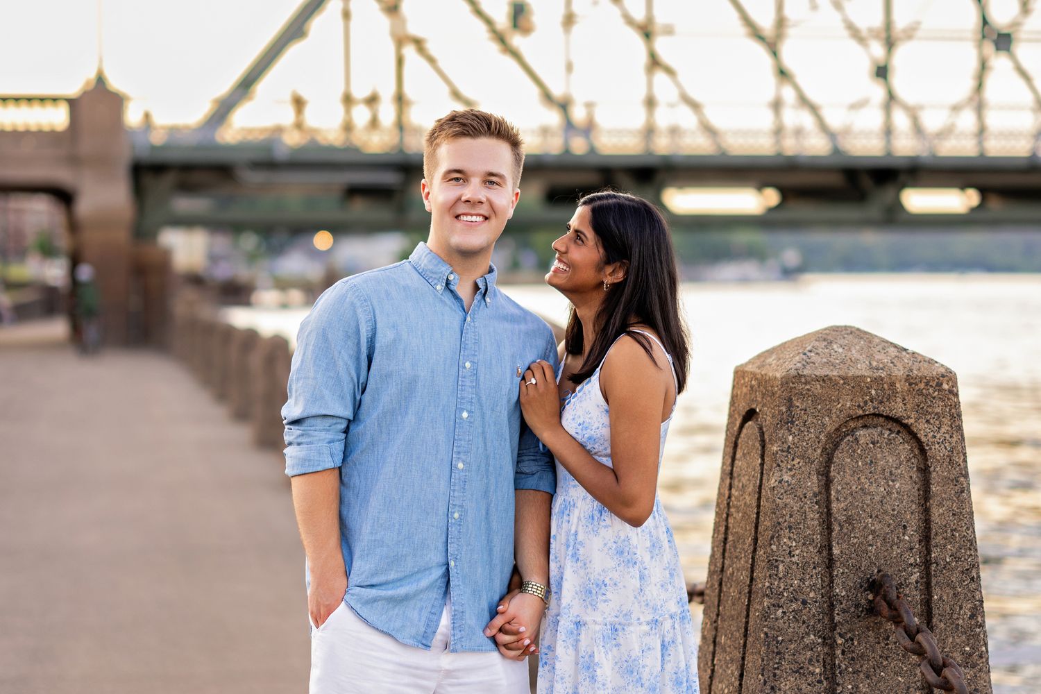 Smiling couple posing together on a waterfront promenade with a historic bridge behind them.