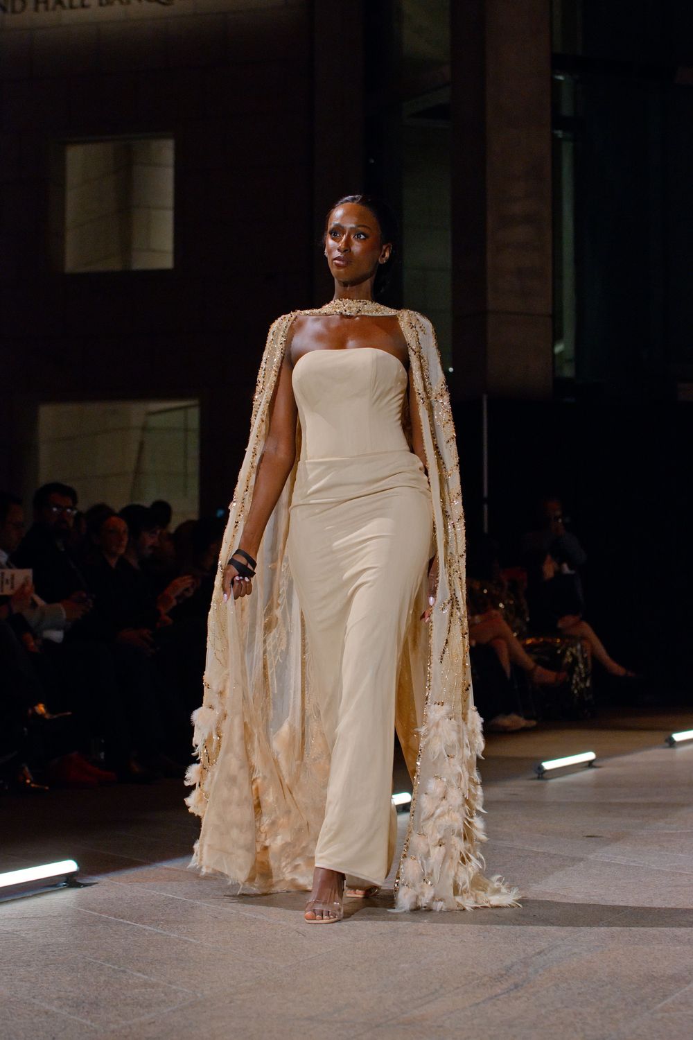 Model walks runway in elegant cream strapless jumpsuit with feather-trimmed cape and gold choker necklace.