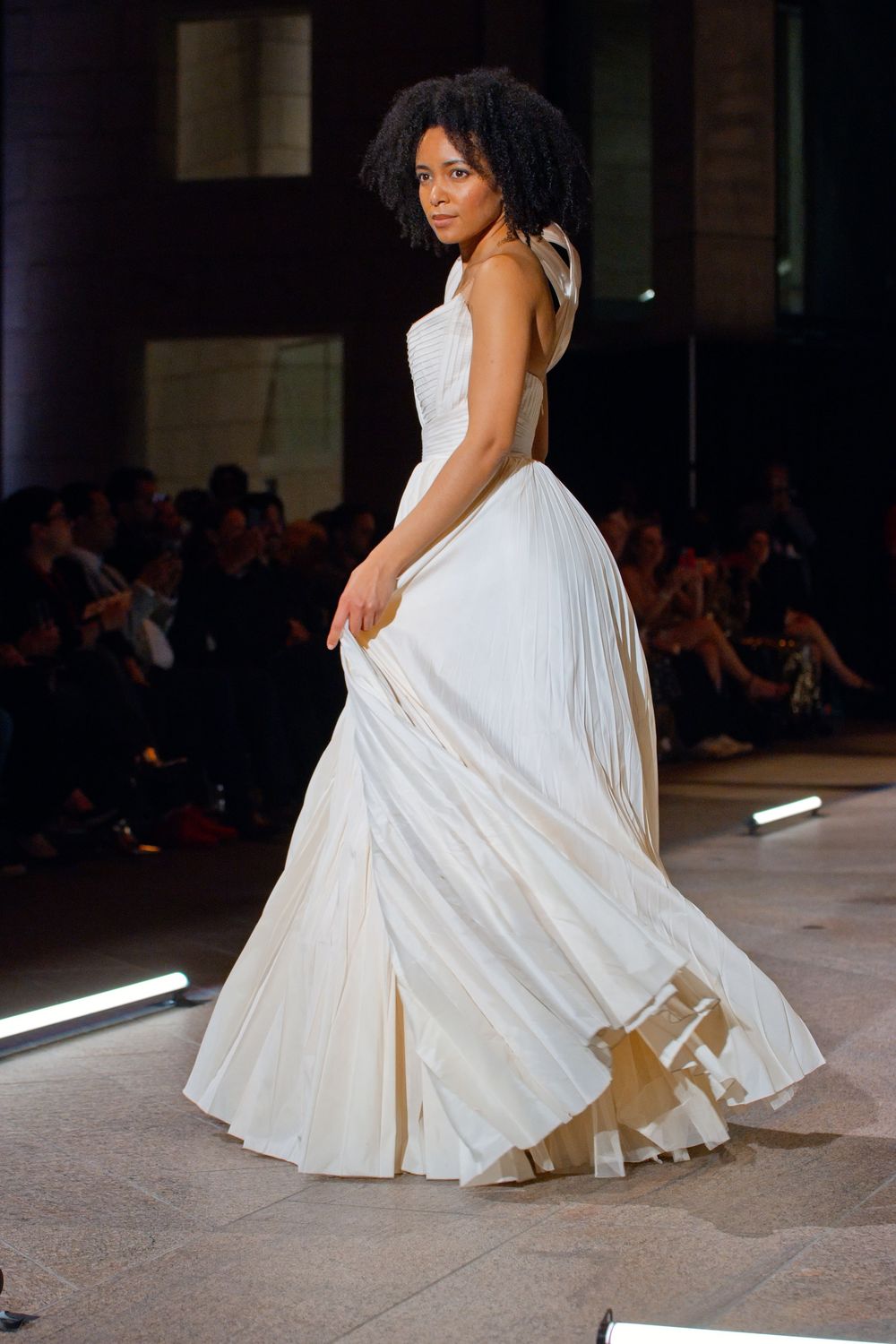 Model walks runway in flowing white pleated bridal gown with bow detail at shoulder during fashion show.