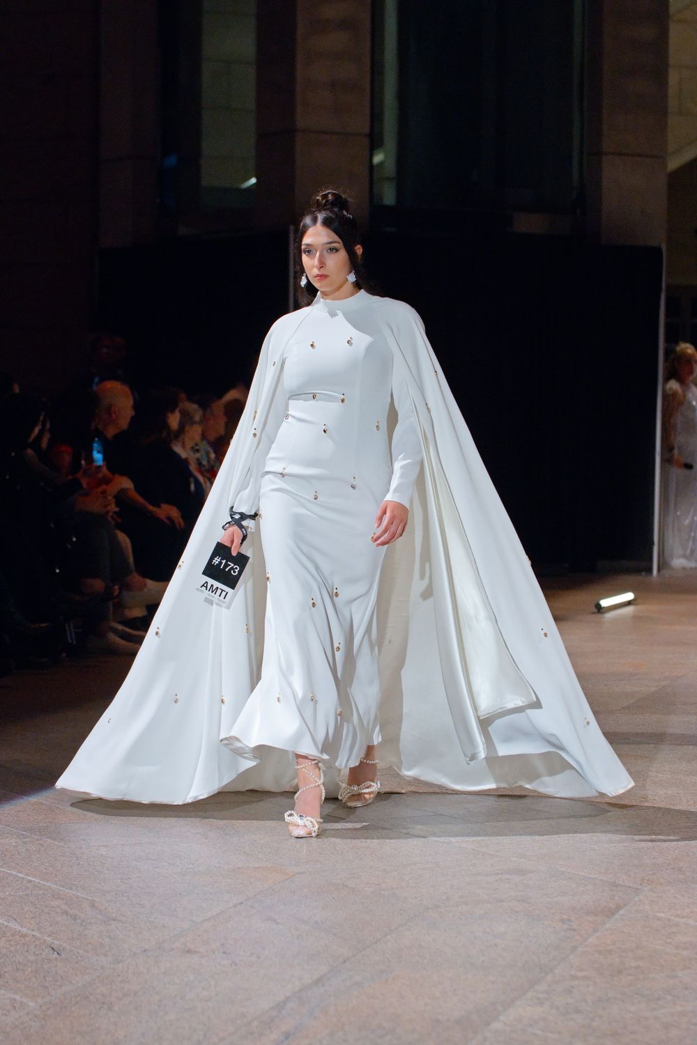 Model walks runway in elegant white cape gown with crystal embellishments at a fashion show.