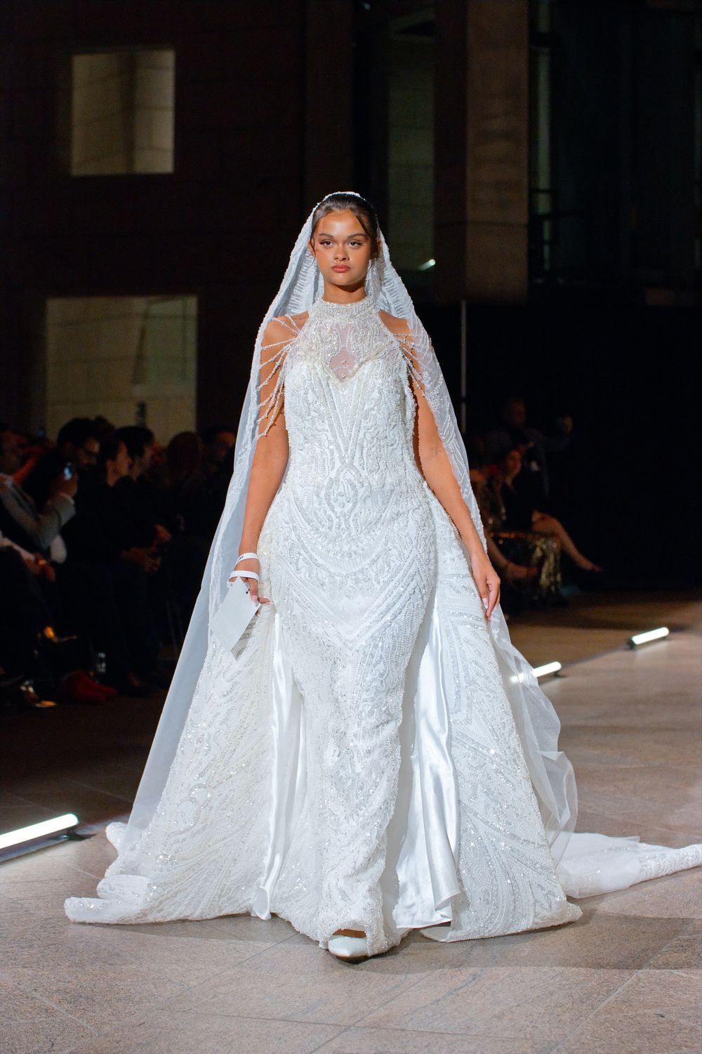 Model showcases ornate beaded white mermaid wedding dress with long veil cape on runway.