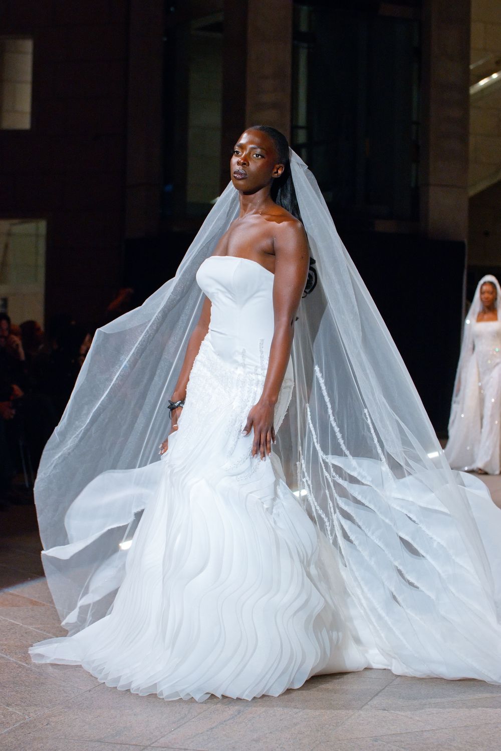 Model walks runway in a strapless white mermaid wedding gown with a flowing cathedral veil.