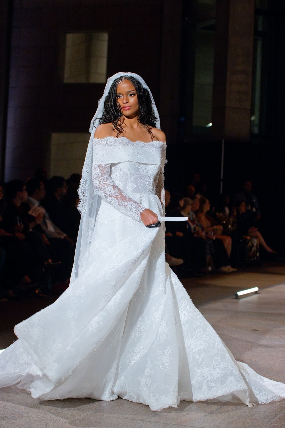 Model walks runway in elegant white lace off-shoulder bridal gown with long sleeves and cathedral veil.