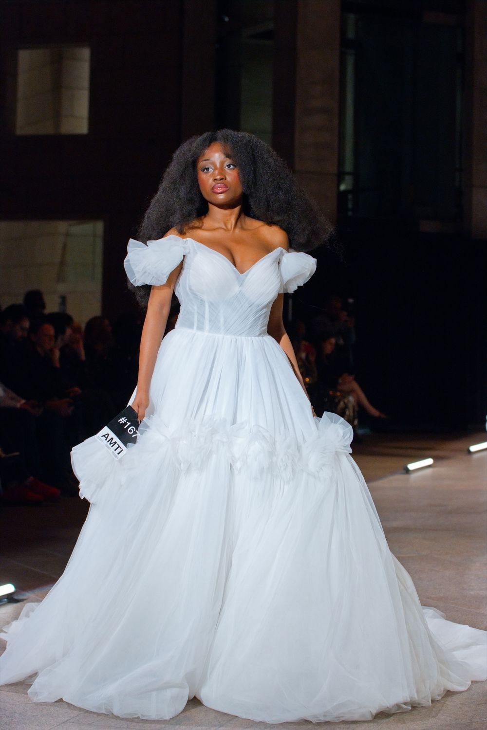 Model showcases a light blue off-shoulder ballgown with ruffled tulle skirt on fashion runway.