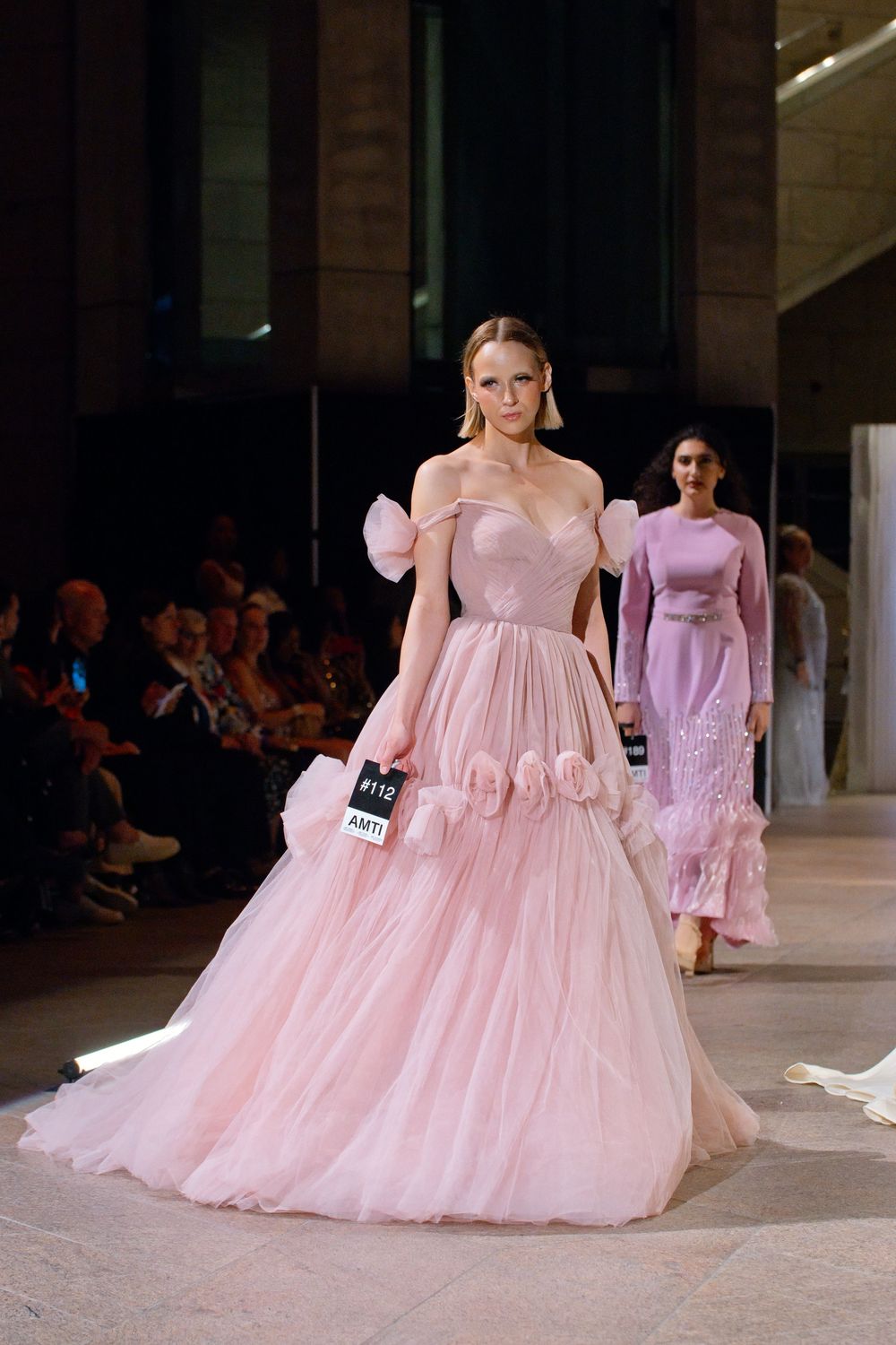 Model showcases elegant blush pink off-shoulder ball gown with floral details and full tulle skirt on fashion runway.