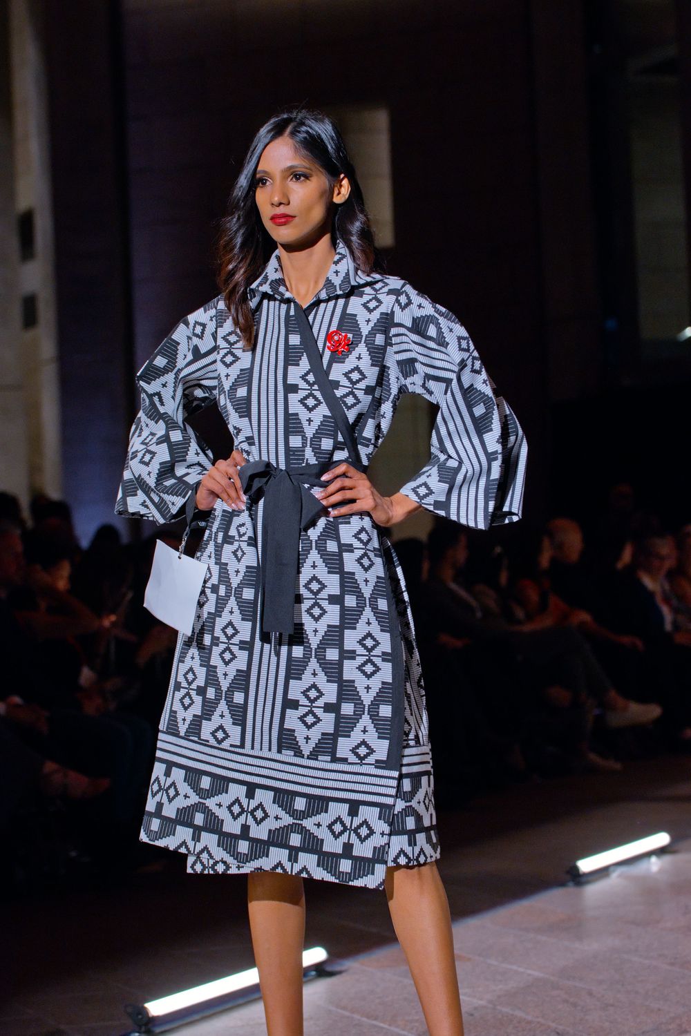 Model walks runway in gray and white geometric print wrap dress with red brooch at a fashion show.