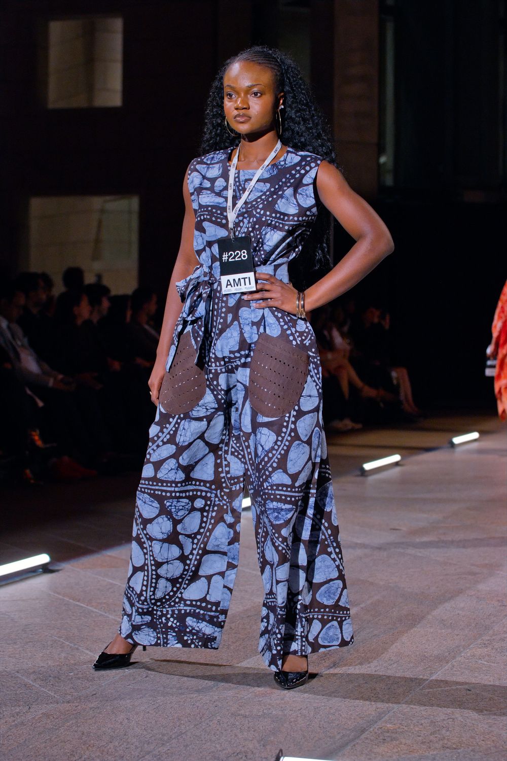 Model walks runway in blue African print jumpsuit with brown crochet pockets at AMTI fashion show.