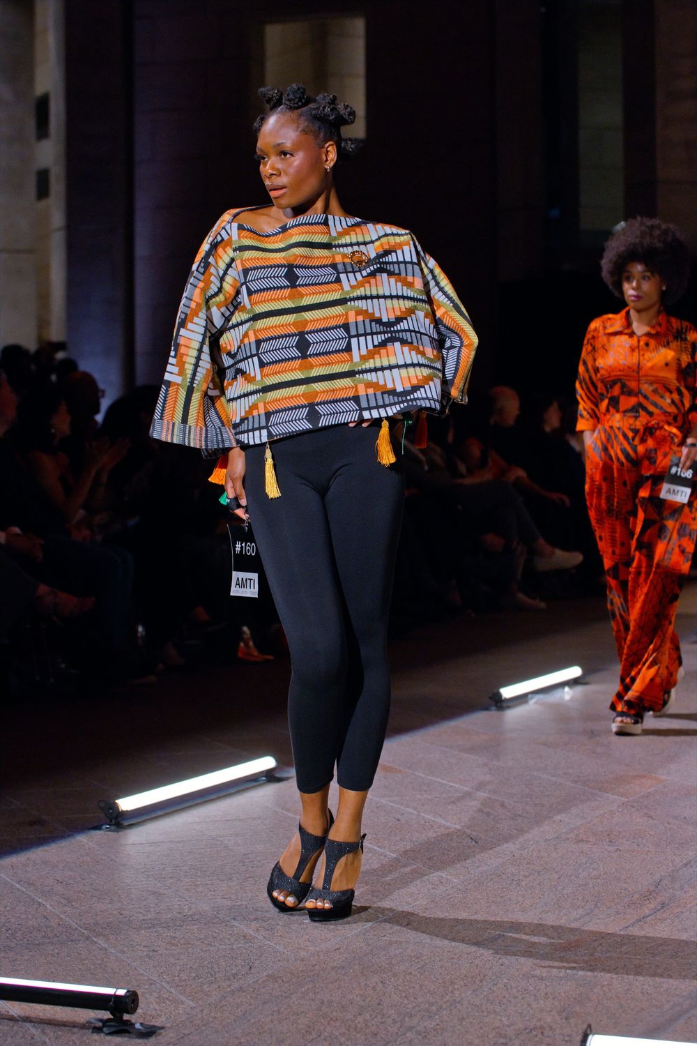 Model walks runway in African kente print top with tassels, black leggings, and heels at a fashion show.