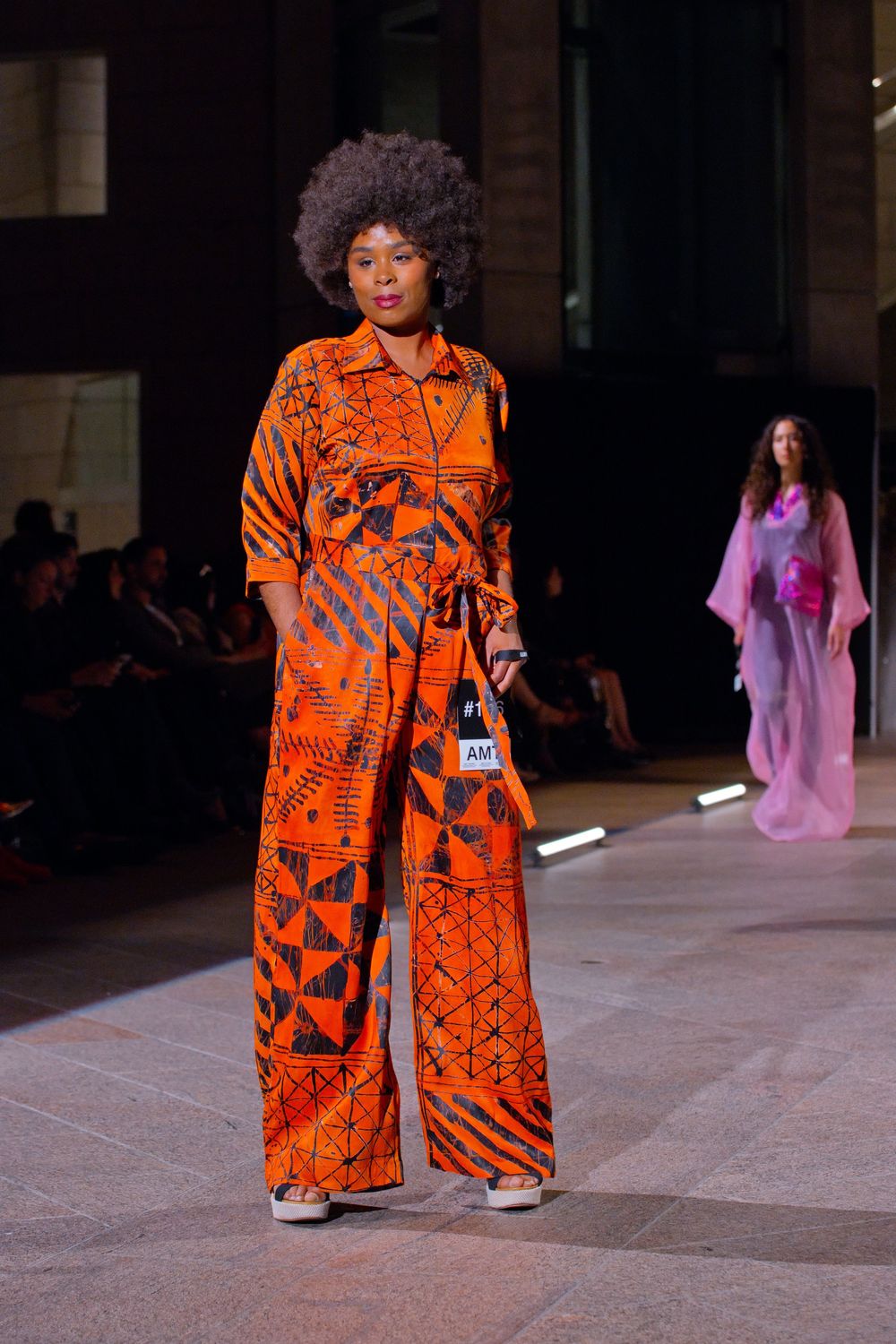Model walks runway in bold orange African print jumpsuit with wide-leg trousers and natural afro hairstyle at fashion show.