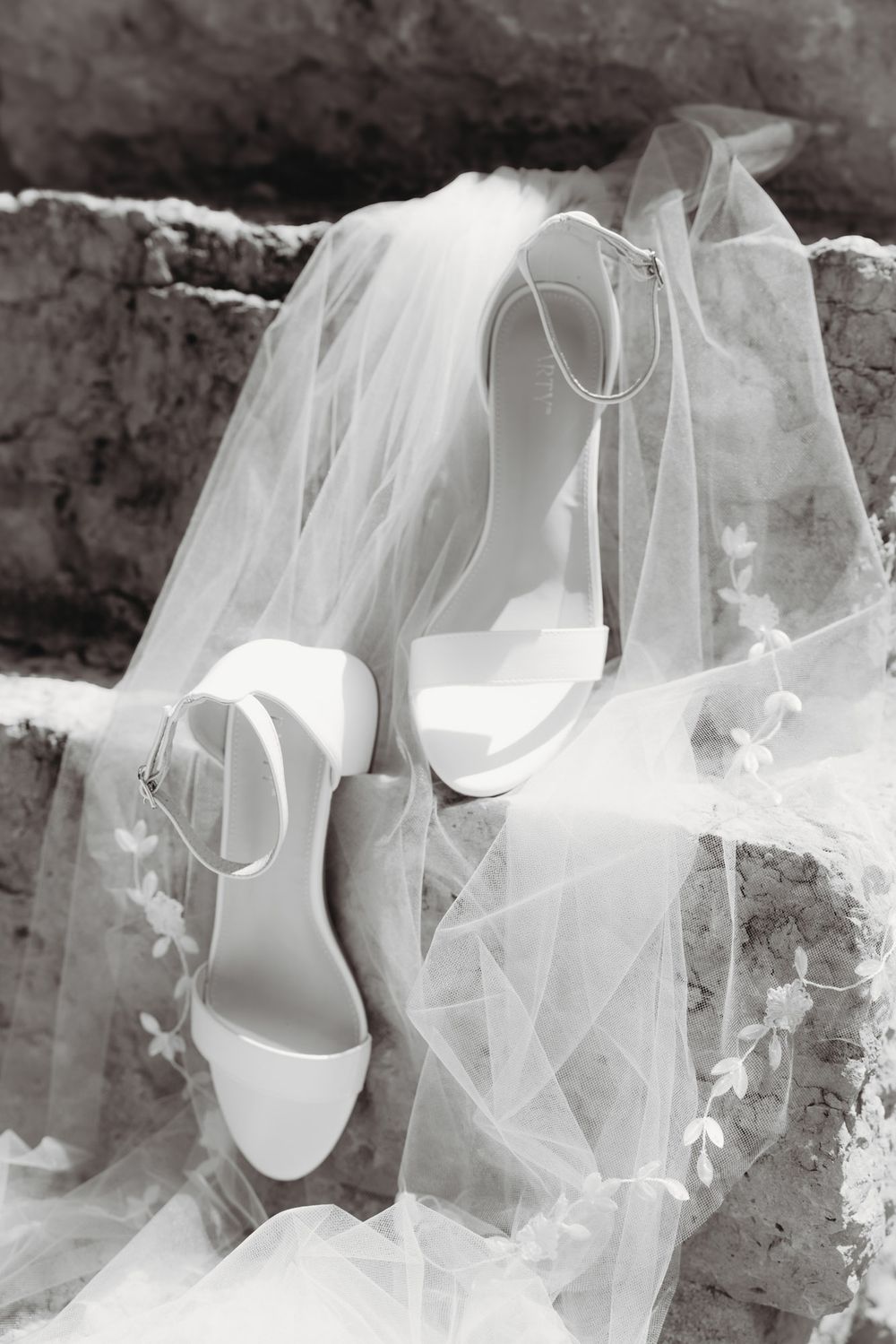 Black and white photo of elegant bridal heels with ankle straps resting on stone steps with a delicate floral veil.