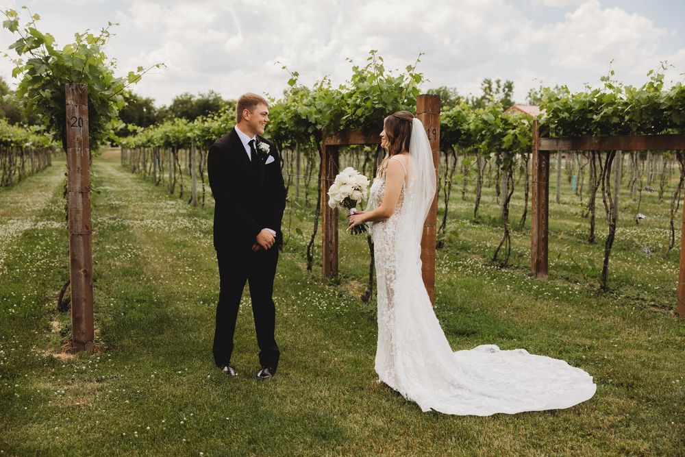 Bride and groom share a first look moment among lush vineyard rows, bride in lace gown holding white bouquet.