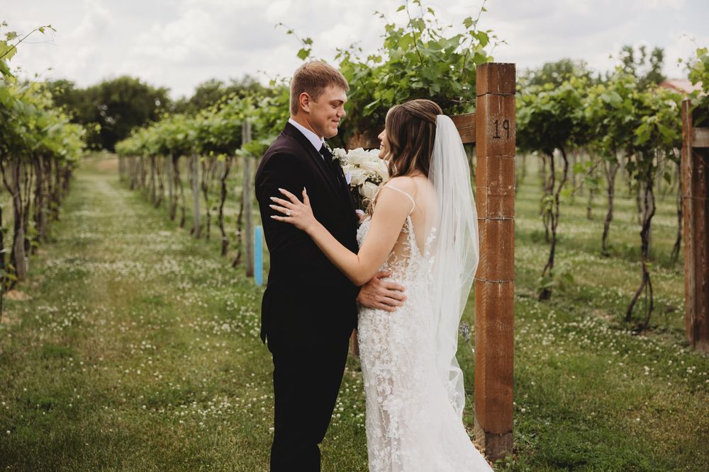Bride and groom share a romantic moment in a lush vineyard, with the bride in a lace gown and veil holding white flowers.