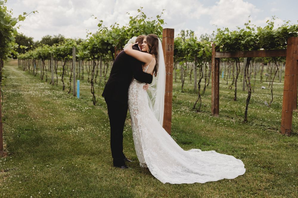 Bride and groom share a romantic kiss in a lush vineyard, bride wearing a flowing white lace gown with veil.