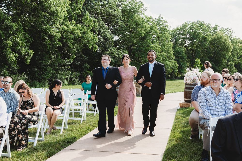 Bride walks down outdoor wedding aisle escorted by two men, guests seated on white chairs, lush green trees in background.